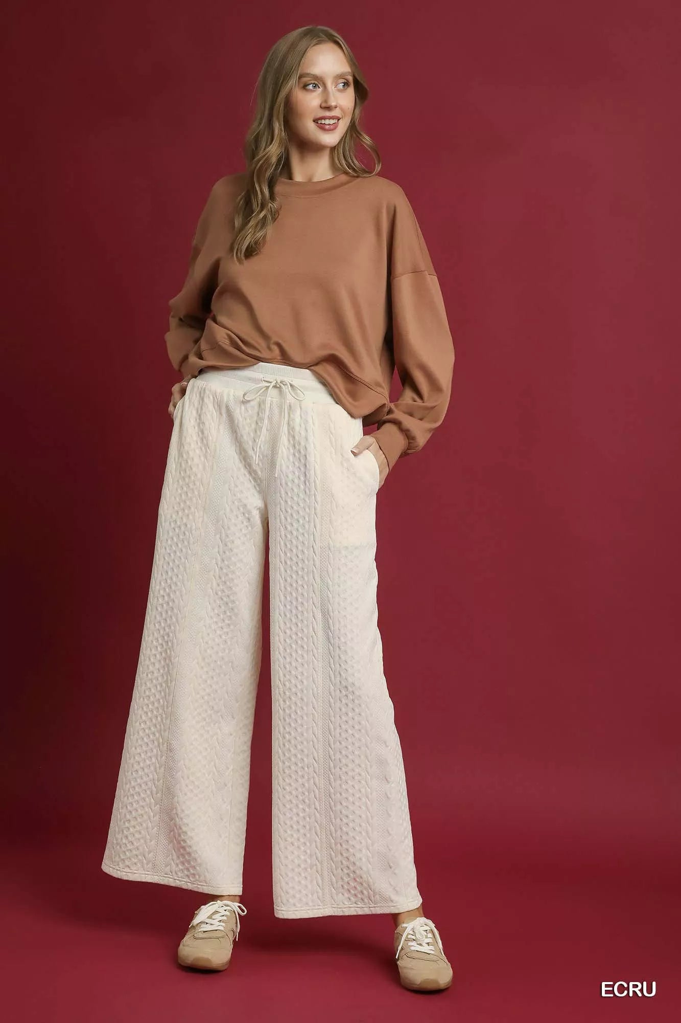 Model poses in cream cable knit textured wide leg pants with drawstring waist on red background.