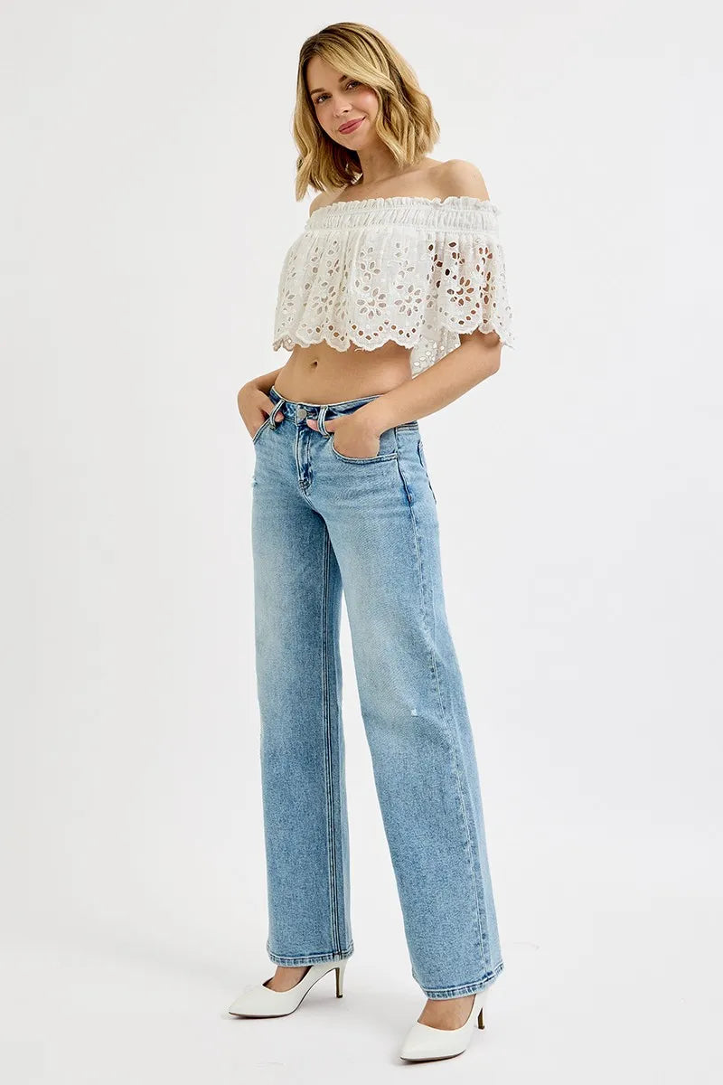Model poses in light wash wide leg flare boyfriend jeans with white lace off-shoulder top and white heels.