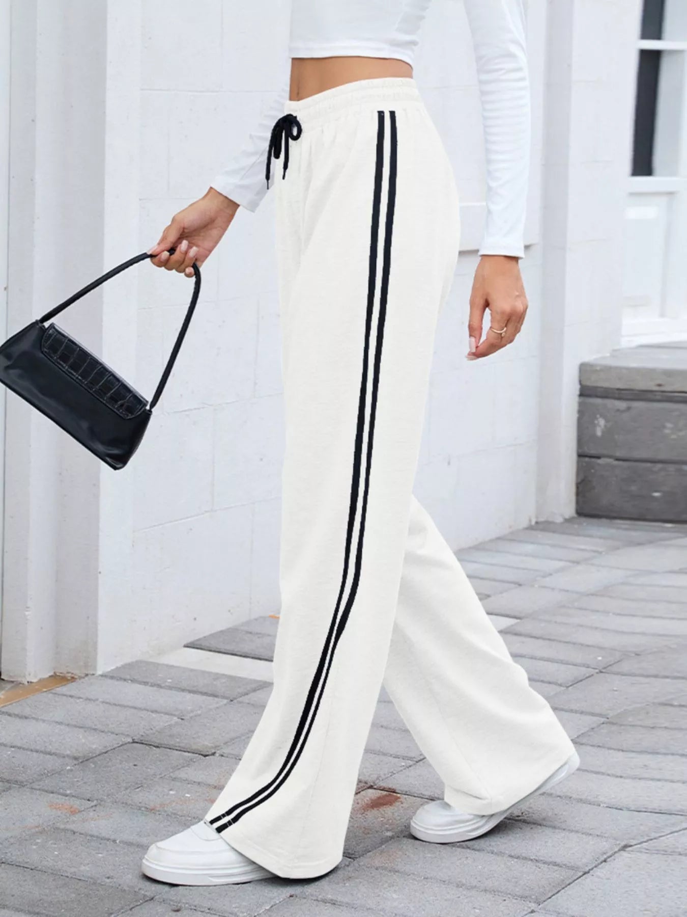 White wide-leg pants with black side stripes, drawstring waist, modeled by a woman holding a black bag.