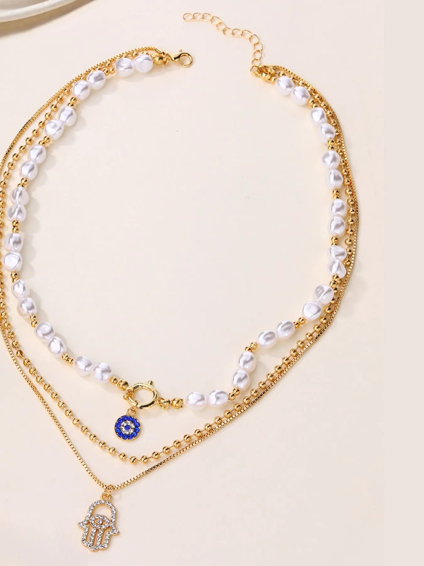 Layered necklace with white pearls, gold-plated chains, blue evil eye, and hamsa pendant.