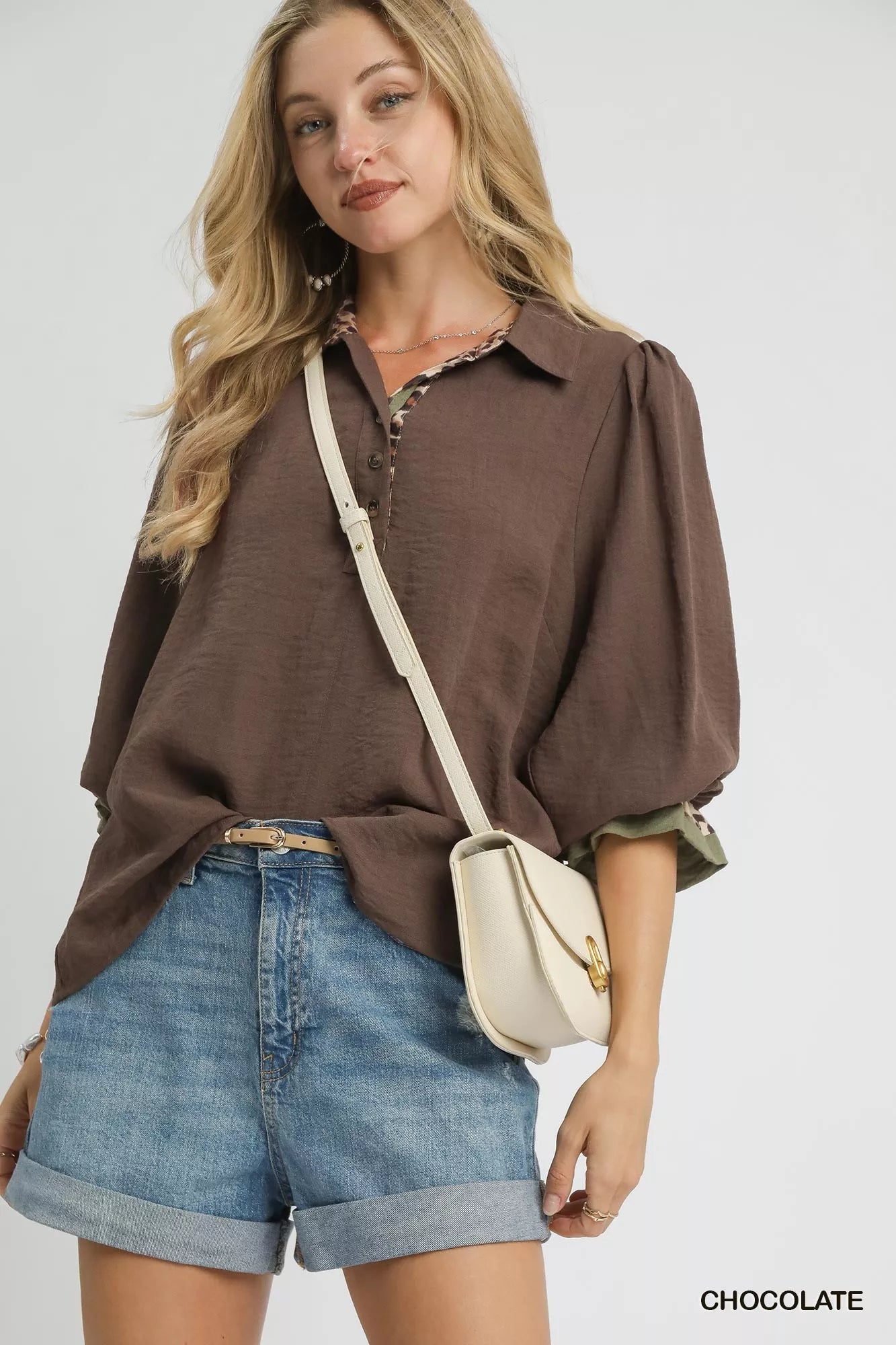Woman wearing a brown half-button blouse with leopard print trim, paired with denim shorts and a white crossbody bag.