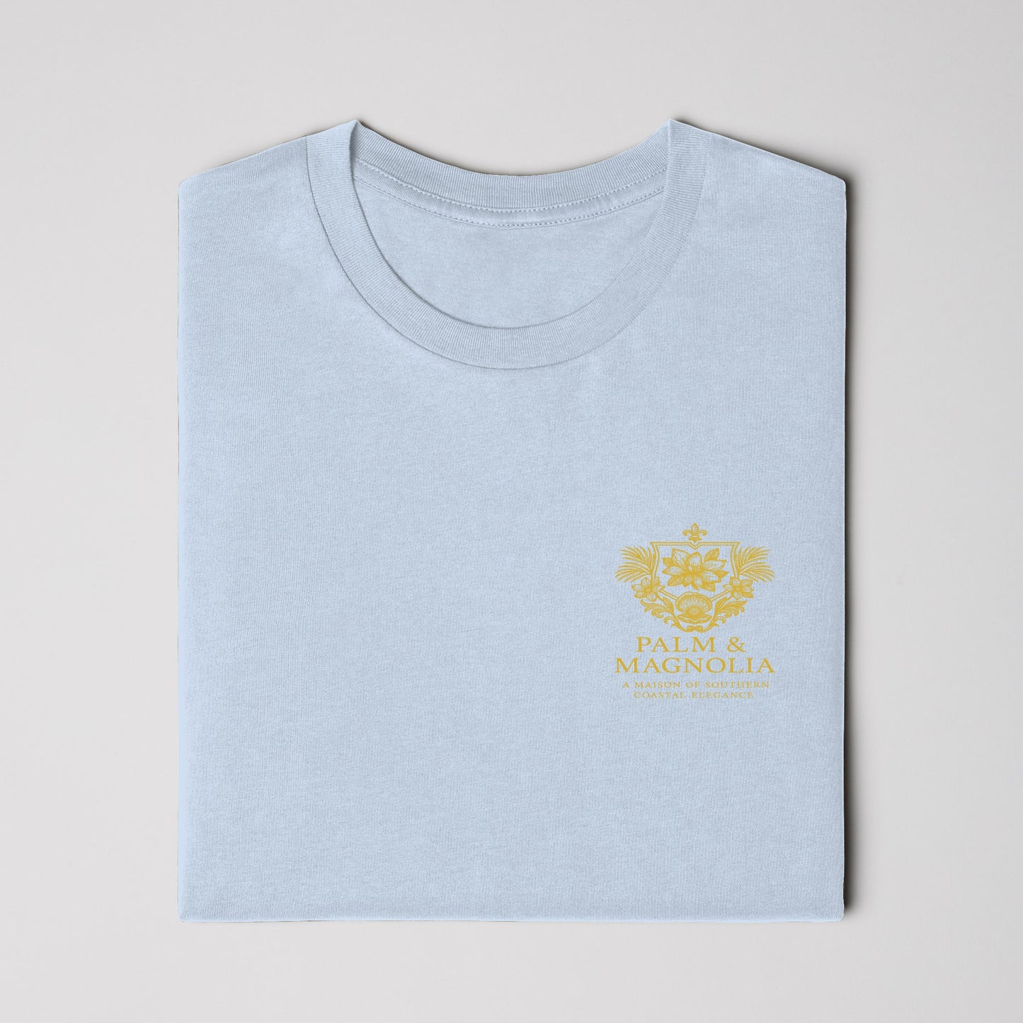 Light blue Bella Canvas T-shirt folded with gold Magnolia Blossom embroidery.