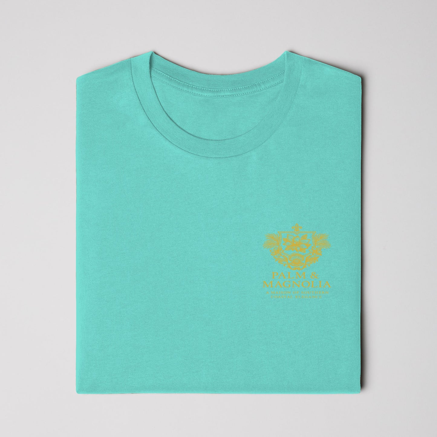 Folded teal Bella Canvas t-shirt with gold embroidered magnolia blossom on chest.