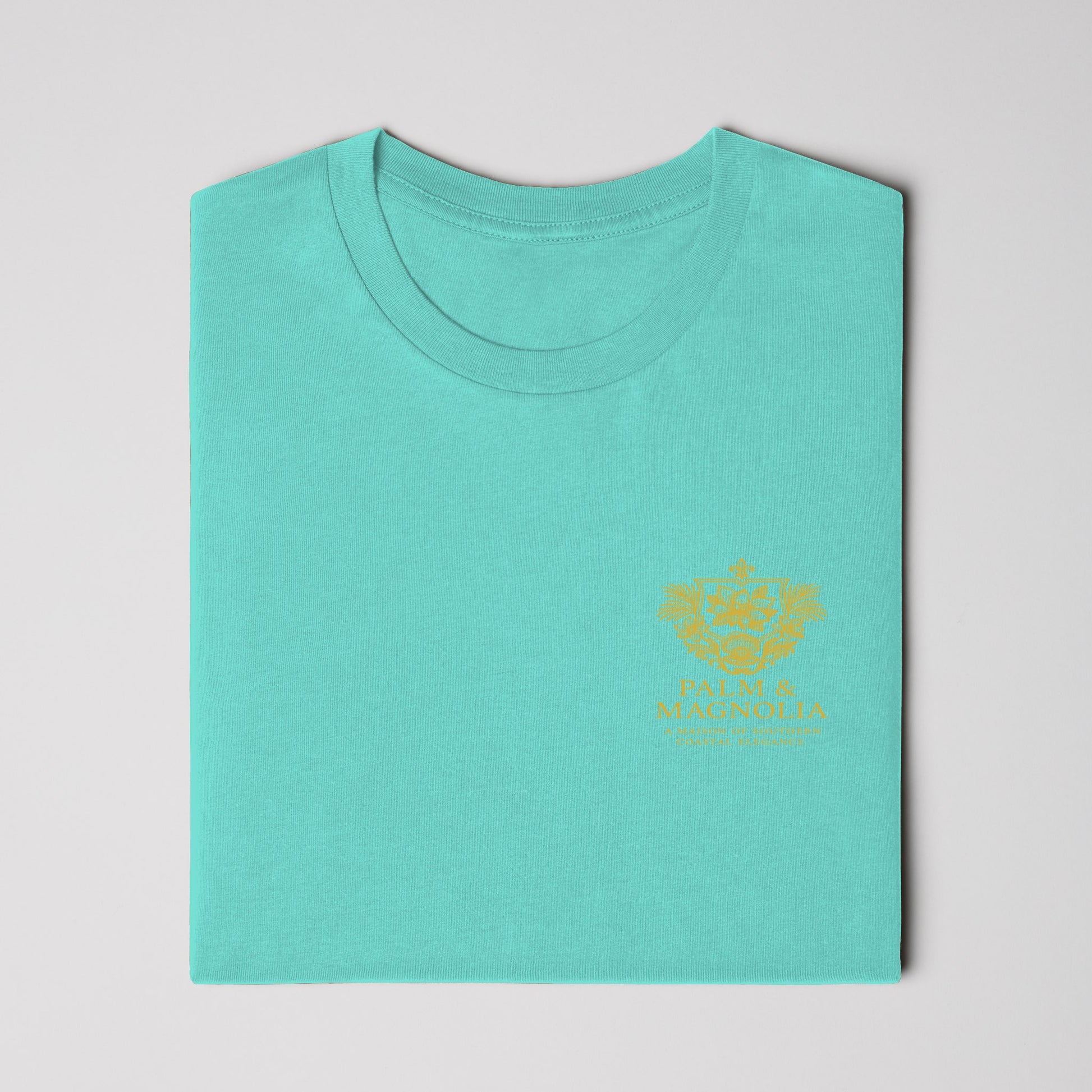 Folded teal Bella Canvas t-shirt with gold embroidered magnolia blossom on chest.