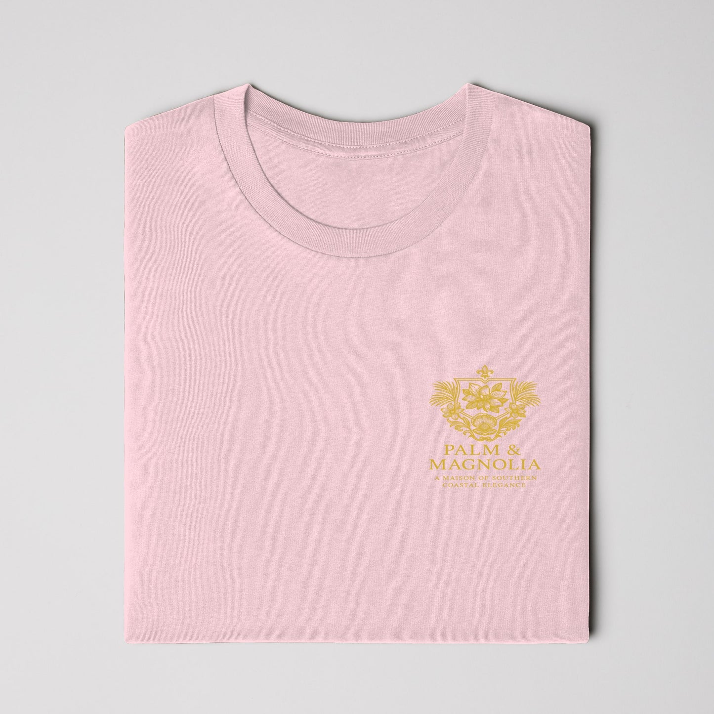 Folded pink Bella Canvas T-shirt with gold Magnolia Blossom embroidery on chest.