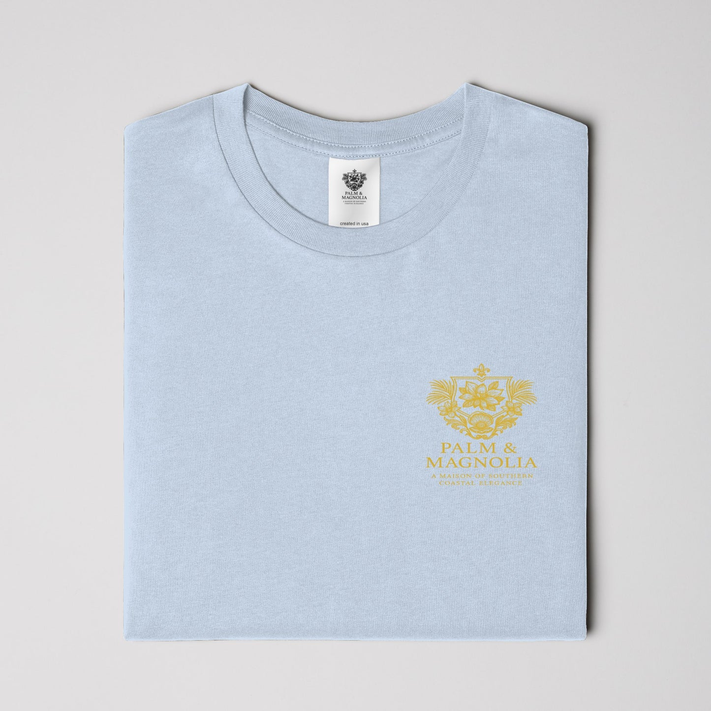 Folded light blue unisex tee with gold Palm & Magnolia logo on chest.