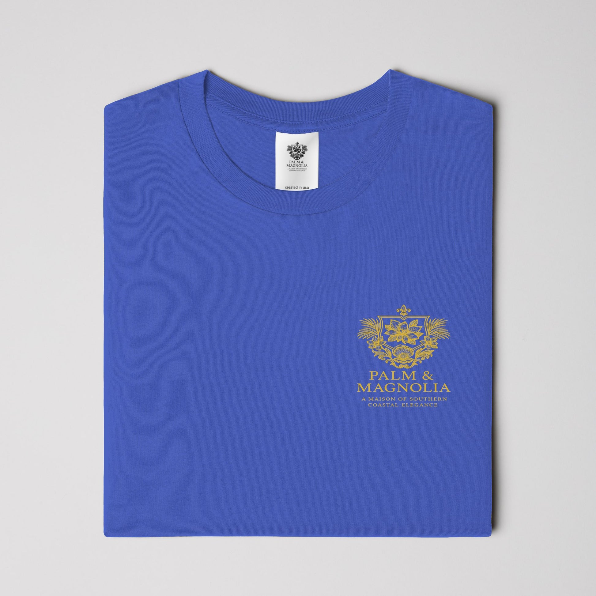 Folded blue unisex tee with gold Palm & Magnolia logo on chest.