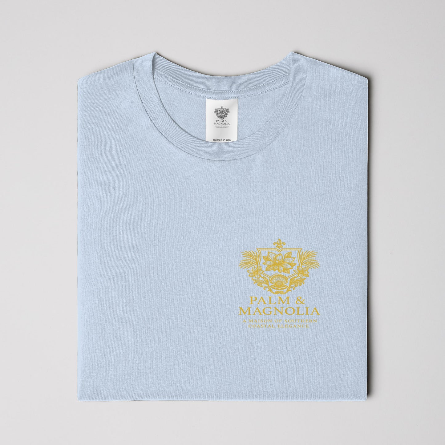 Light blue unisex ring-spun cotton tee folded, featuring gold Palm & Magnolia crest.