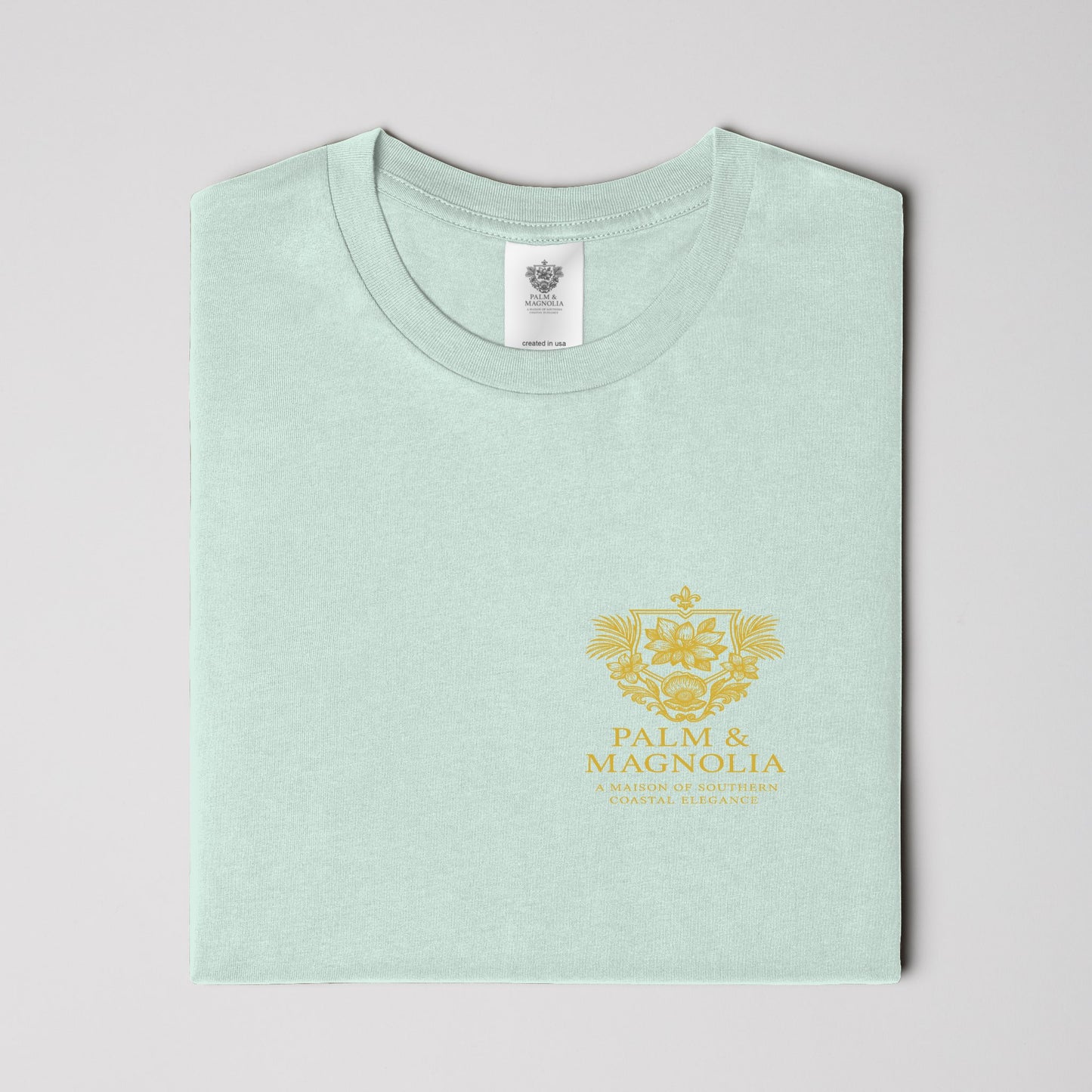 Folded light green unisex tee with gold Palm & Magnolia crest emblem.