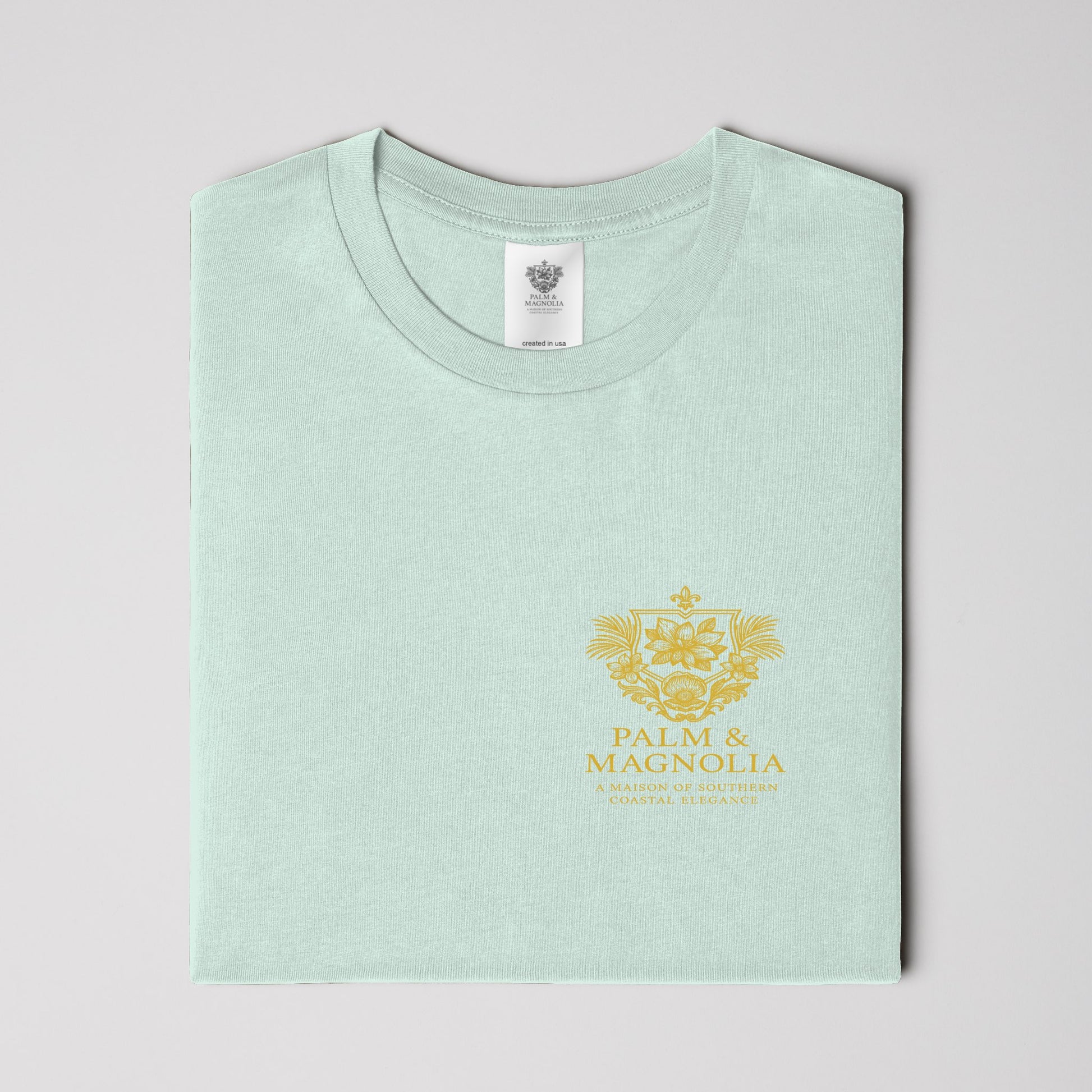 Folded light green unisex tee with gold Palm & Magnolia crest emblem.