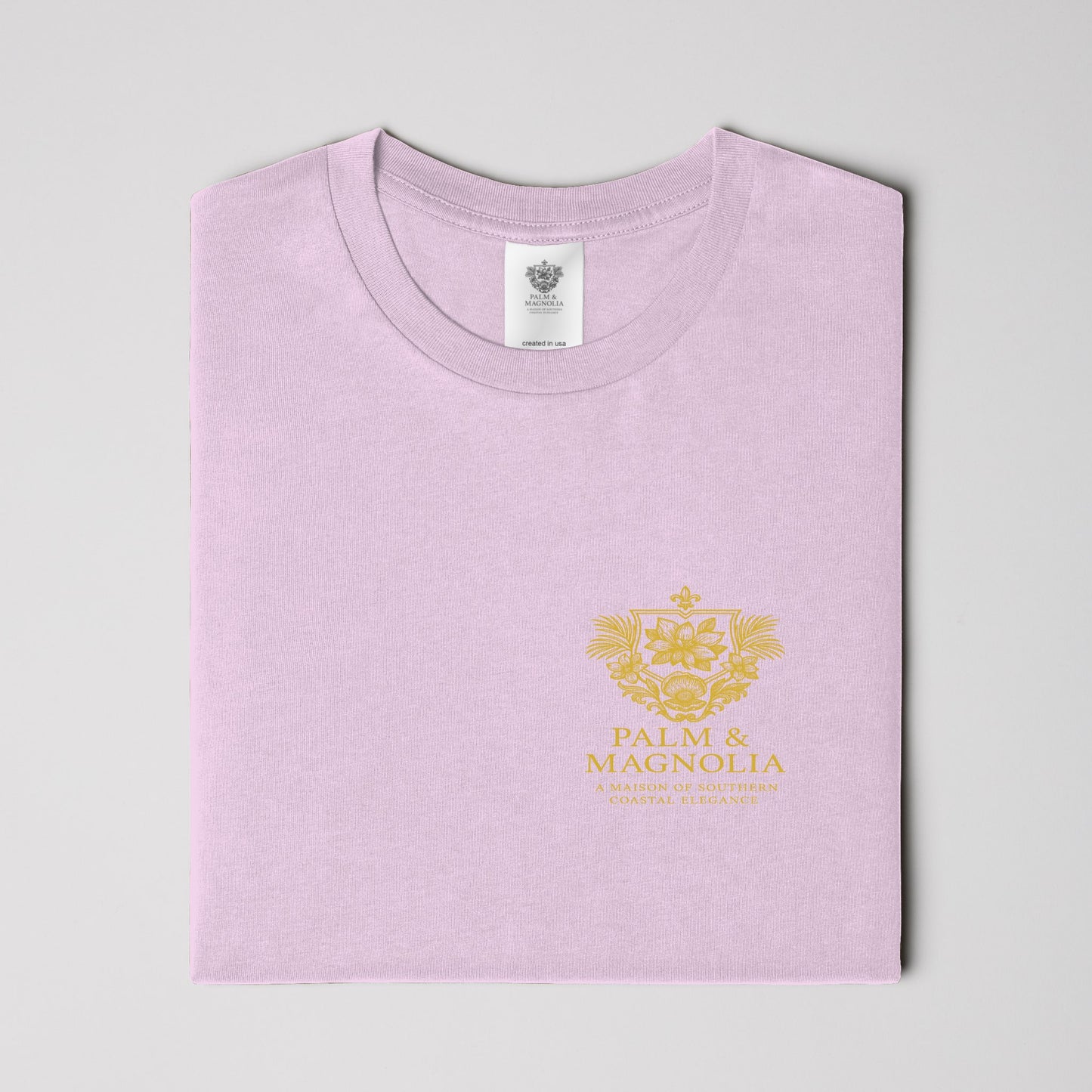 Folded light pink unisex tee with gold Palm & Magnolia crest on chest.