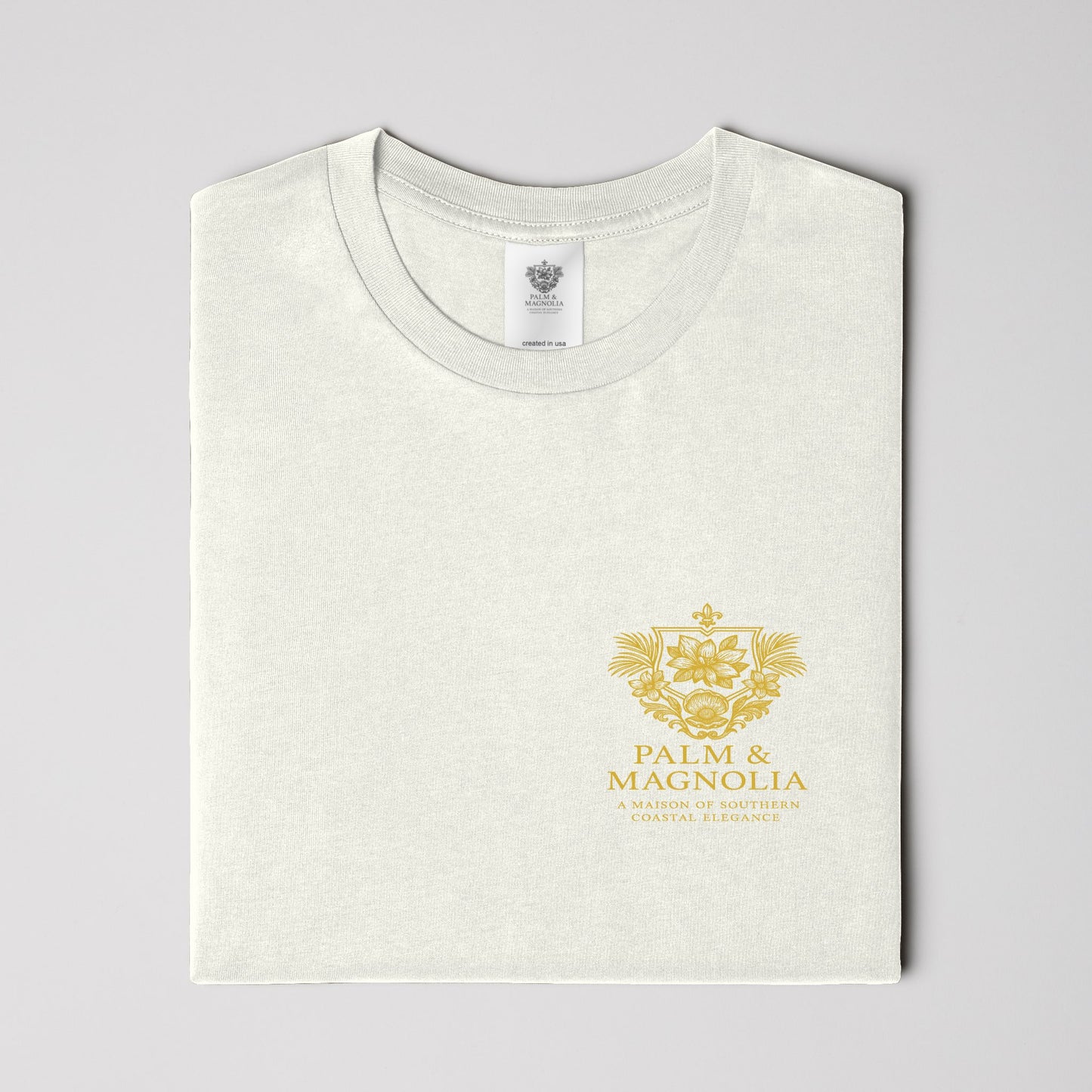 Folded white Palm & Magnolia Southern Charm Tee with gold crest on chest.