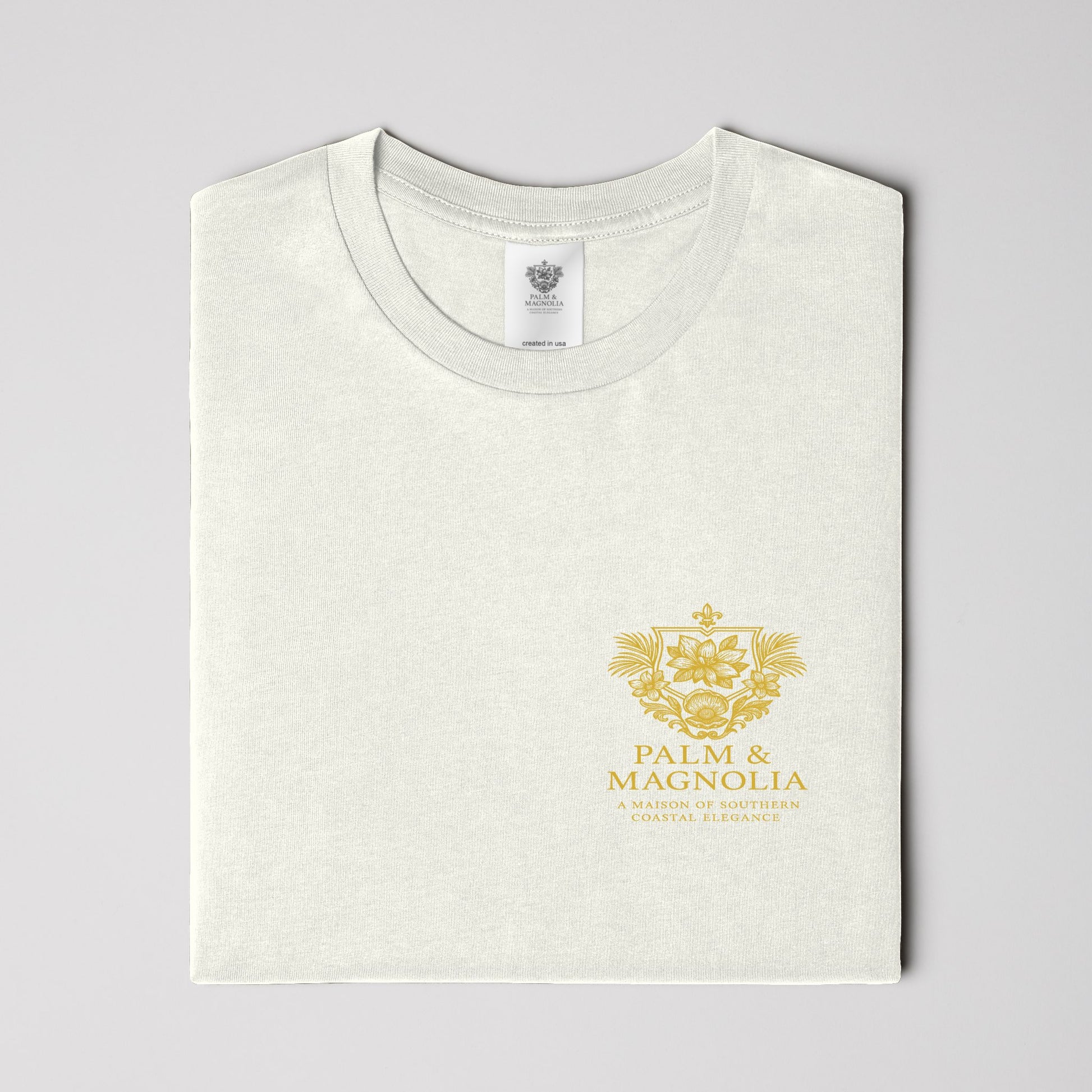 Folded white Palm & Magnolia Southern Charm Tee with gold crest on chest.
