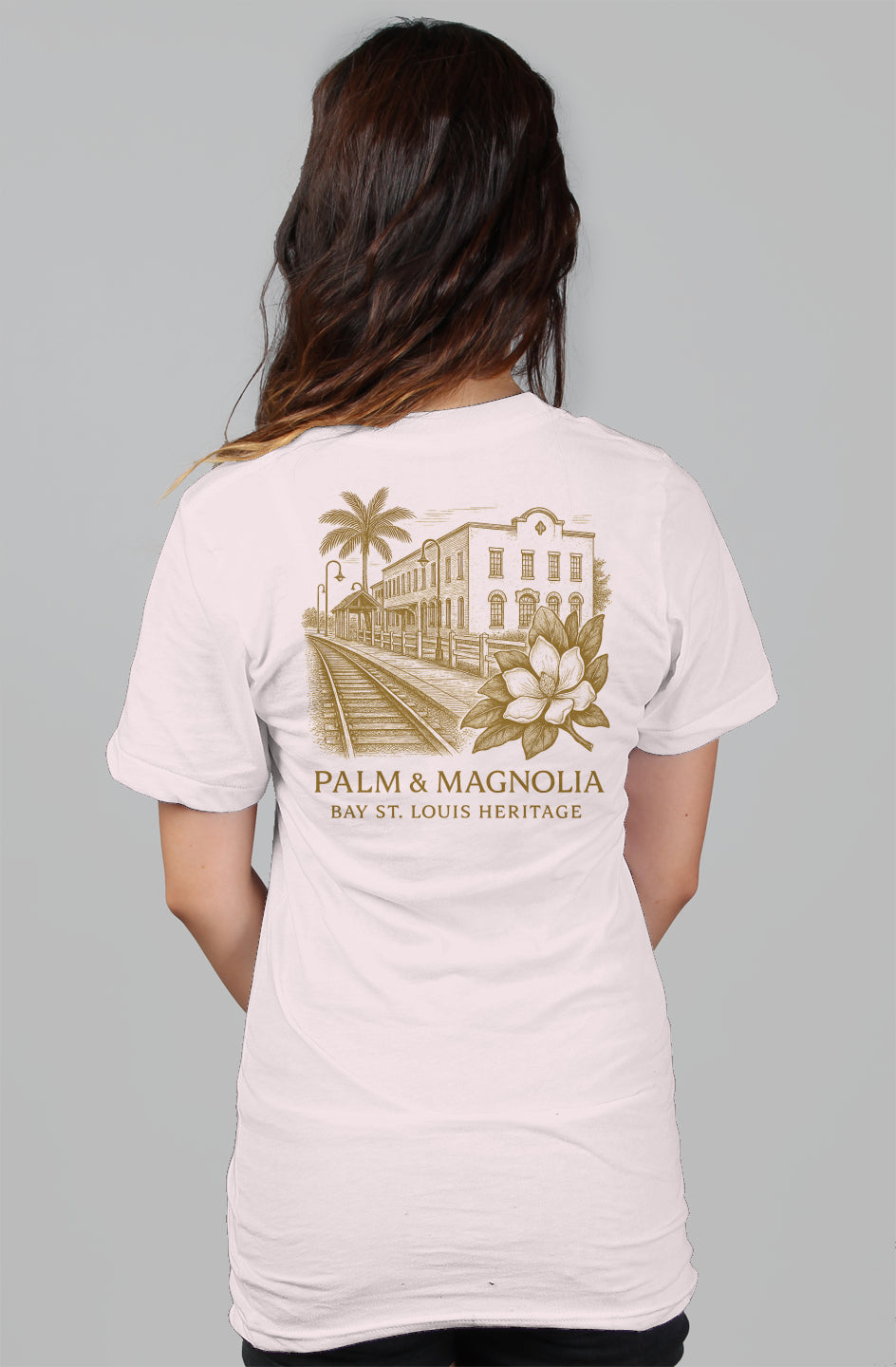 Back view of a white relaxed women's tee featuring a gold Palm & Magnolia Bay St. Louis crest with palm tree, building, and magnolia.