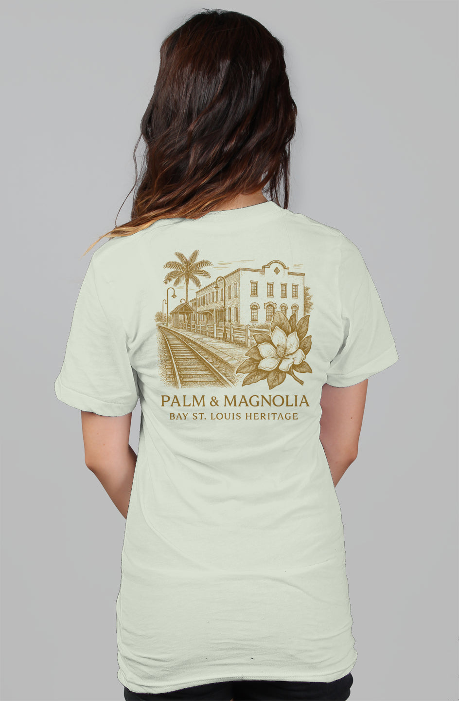Back view of a relaxed women's light gray tee with gold Palm & Magnolia Bay St. Louis crest featuring a train, building, palm tree, and magnolia.