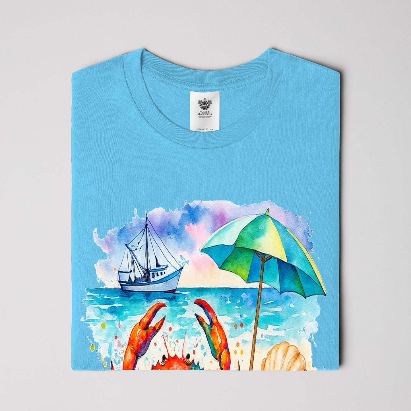 Folded light blue unisex cotton T-shirt with watercolor coastal scene of crab, boat, beach umbrella, and ocean.