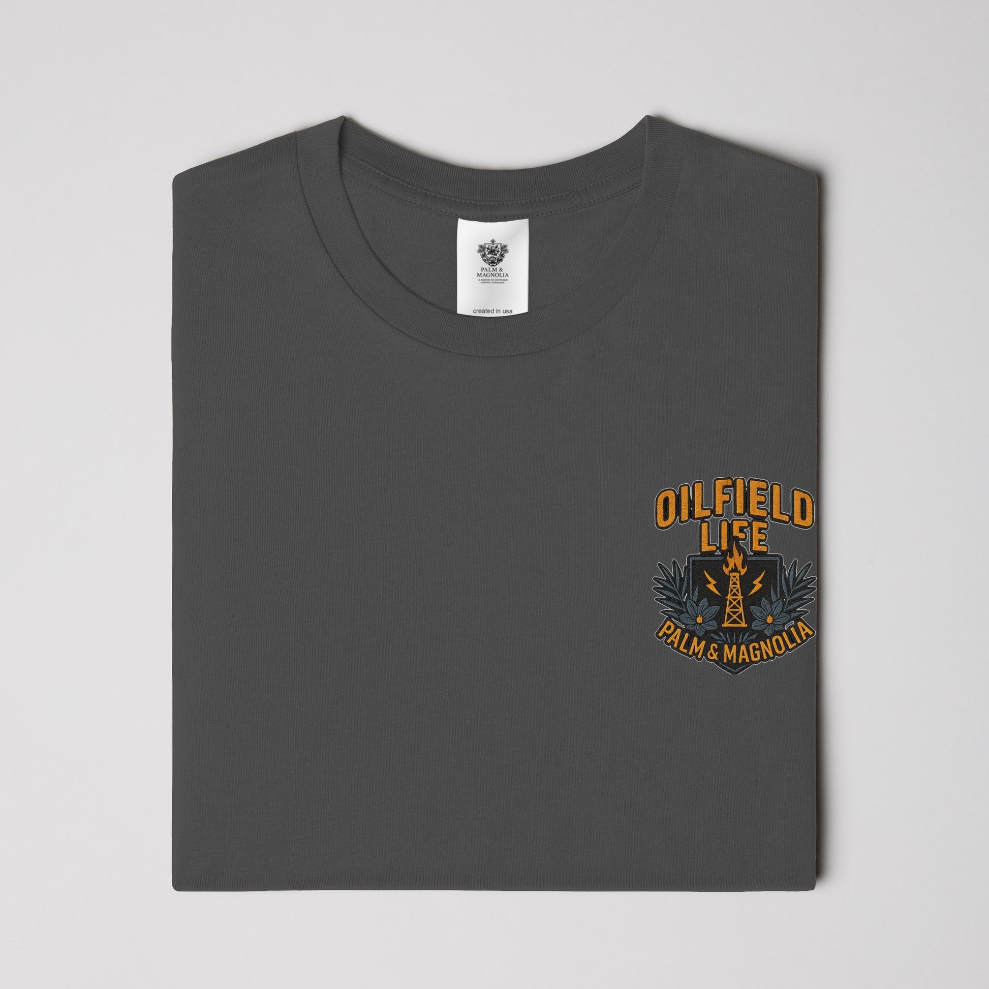 Folded gray heavyweight cotton t-shirt with orange "Oilfield Life Palm & Magnolia" graphic on chest.