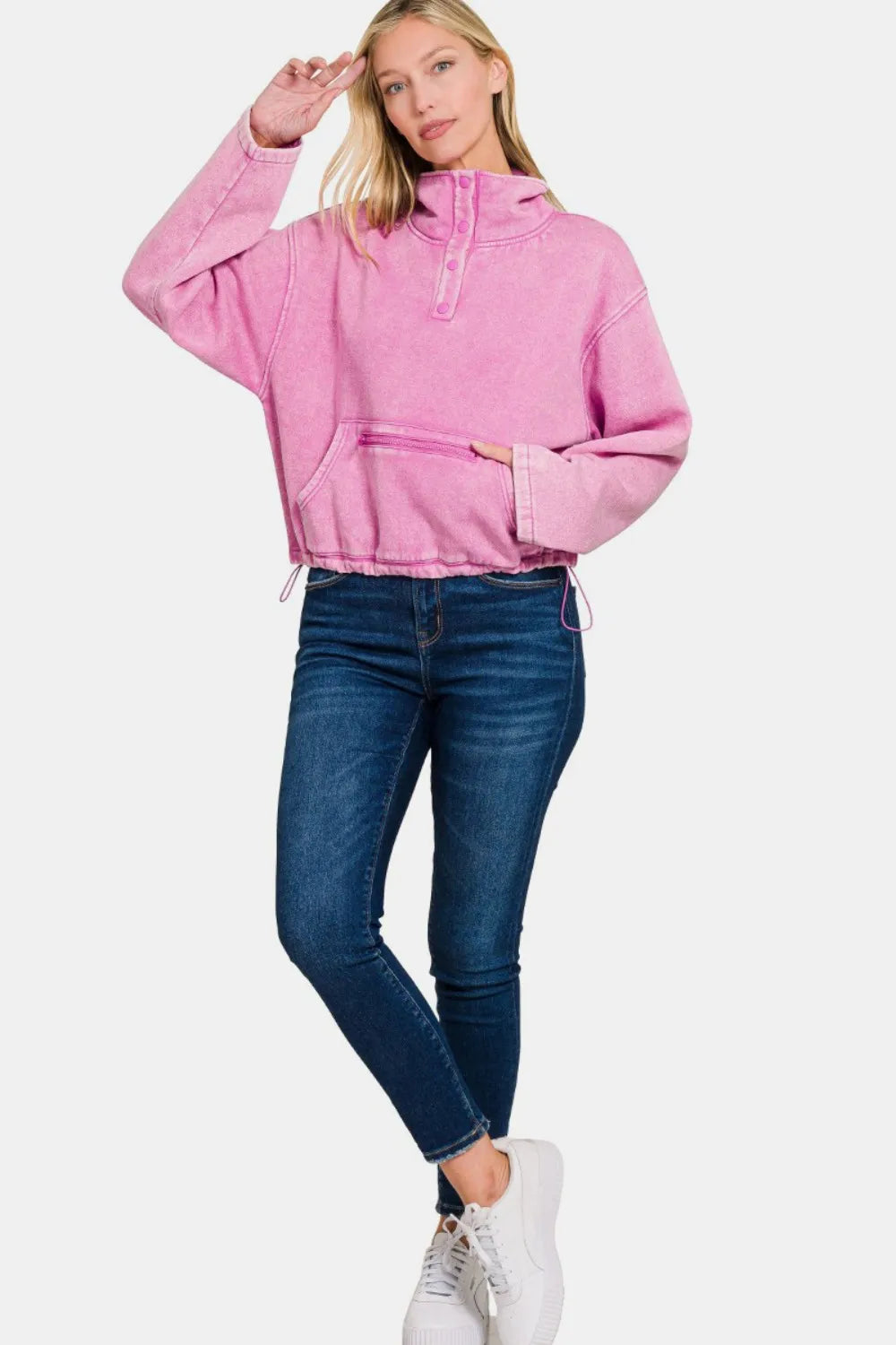 Model poses in pink acid wash fleece half snap sweatshirt with kangaroo pocket and drawstring hood, paired with jeans and white sneakers.