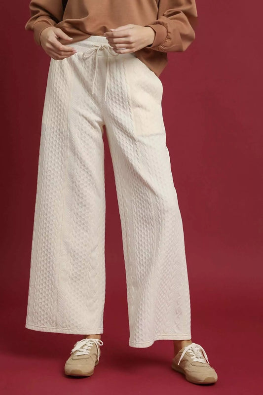White cable knit textured wide leg pants with drawstring waist, modeled on a red background.