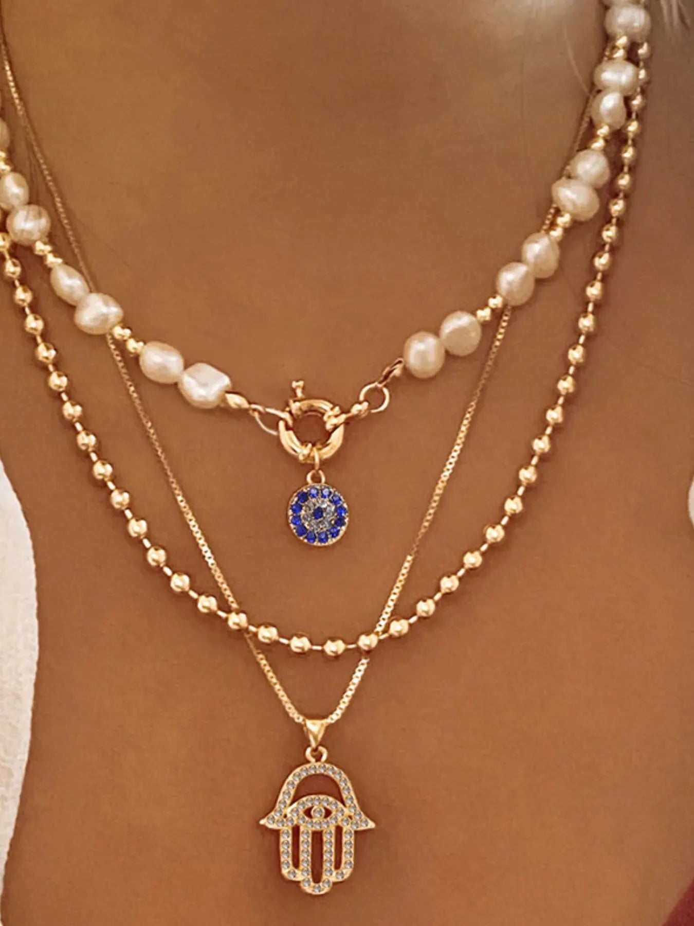 Layered necklace with pearl strands, blue evil eye charm, and gold-plated hamsa pendant.