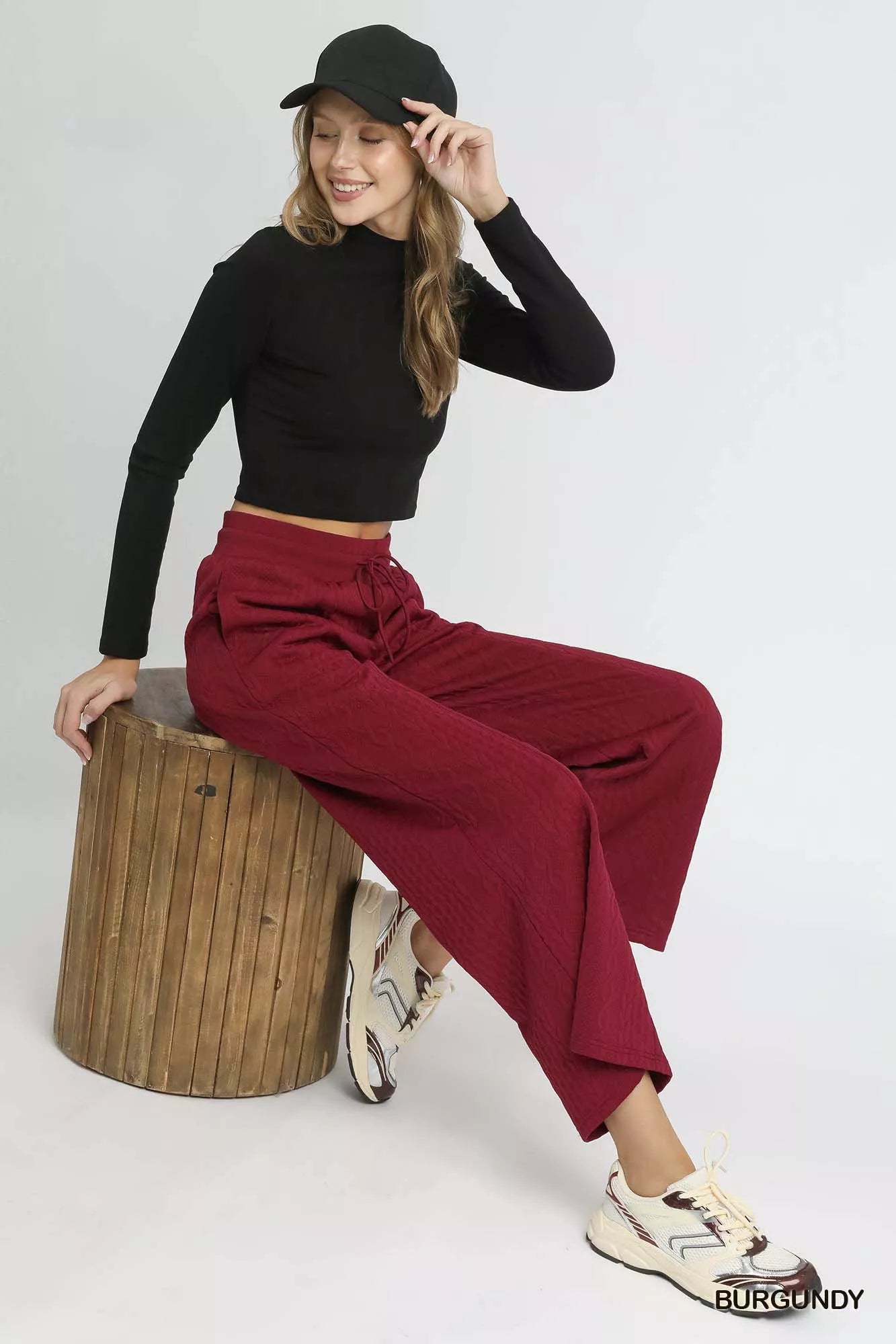 Model wearing black turtleneck, black cap, and burgundy cable knit textured wide leg pants with drawstring, sitting on wooden stool with white sneakers.