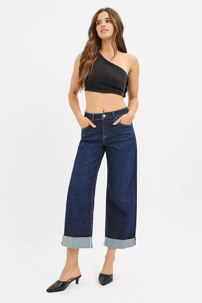 Model poses in RISEN mid-rise ankle wide roll-up jeans with black one-shoulder top and black heels.