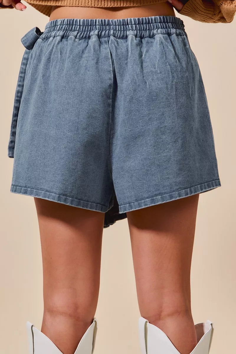 Rear view of SO ME wrap braided trim denim skort with tie detail and white boots.