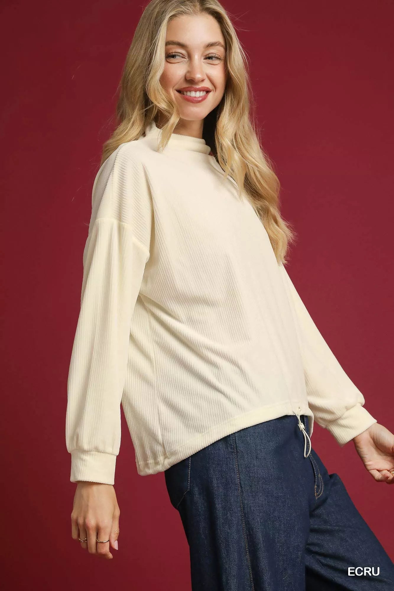 Woman modeling cream ribbed knit top with drawstring hem and long sleeves.