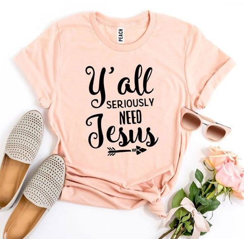 Pink Bella Canvas cotton t-shirt with "Y'all Seriously Need Jesus" text, flat-laid with espadrilles, sunglasses, and roses.