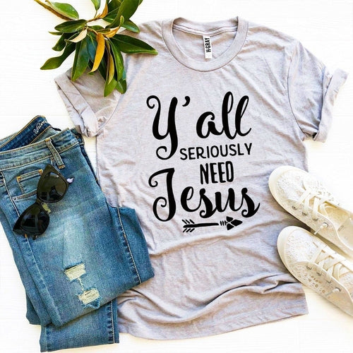 Gray Bella Canvas cotton t-shirt with "Y'all Seriously Need Jesus" text and arrow, styled with jeans, sneakers, and greenery.