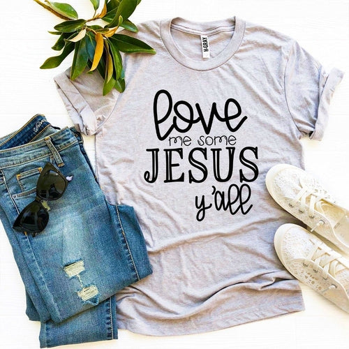 Gray Bella Canvas cotton t-shirt with "Love Me Some Jesus Y'all" text, styled with jeans, sneakers, and greenery.