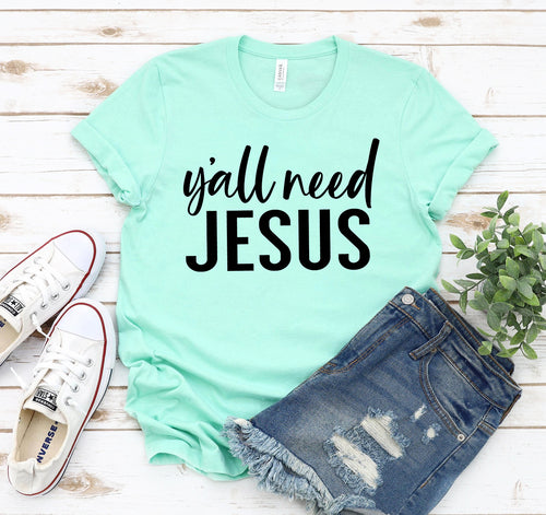 Light green unisex Bella Canvas T-shirt with "Y'all Need Jesus" text, on wood with white sneakers and denim shorts.