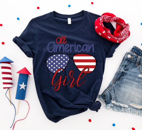 Navy Bella Canvas cotton t-shirt with "All American Girl" text and American flag sunglasses graphic, styled with red bandana and denim shorts on white background.