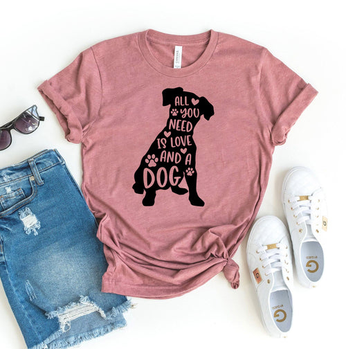 Pink short-sleeve T-shirt with black silhouette of a dog and text "All You Need Is Love And A Dog," styled with distressed shorts and white sneakers.