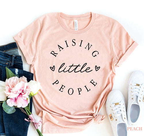 Pink Bella Canvas cotton t-shirt with "Raising Little People" text, styled with jeans, white sneakers, and pink flowers.