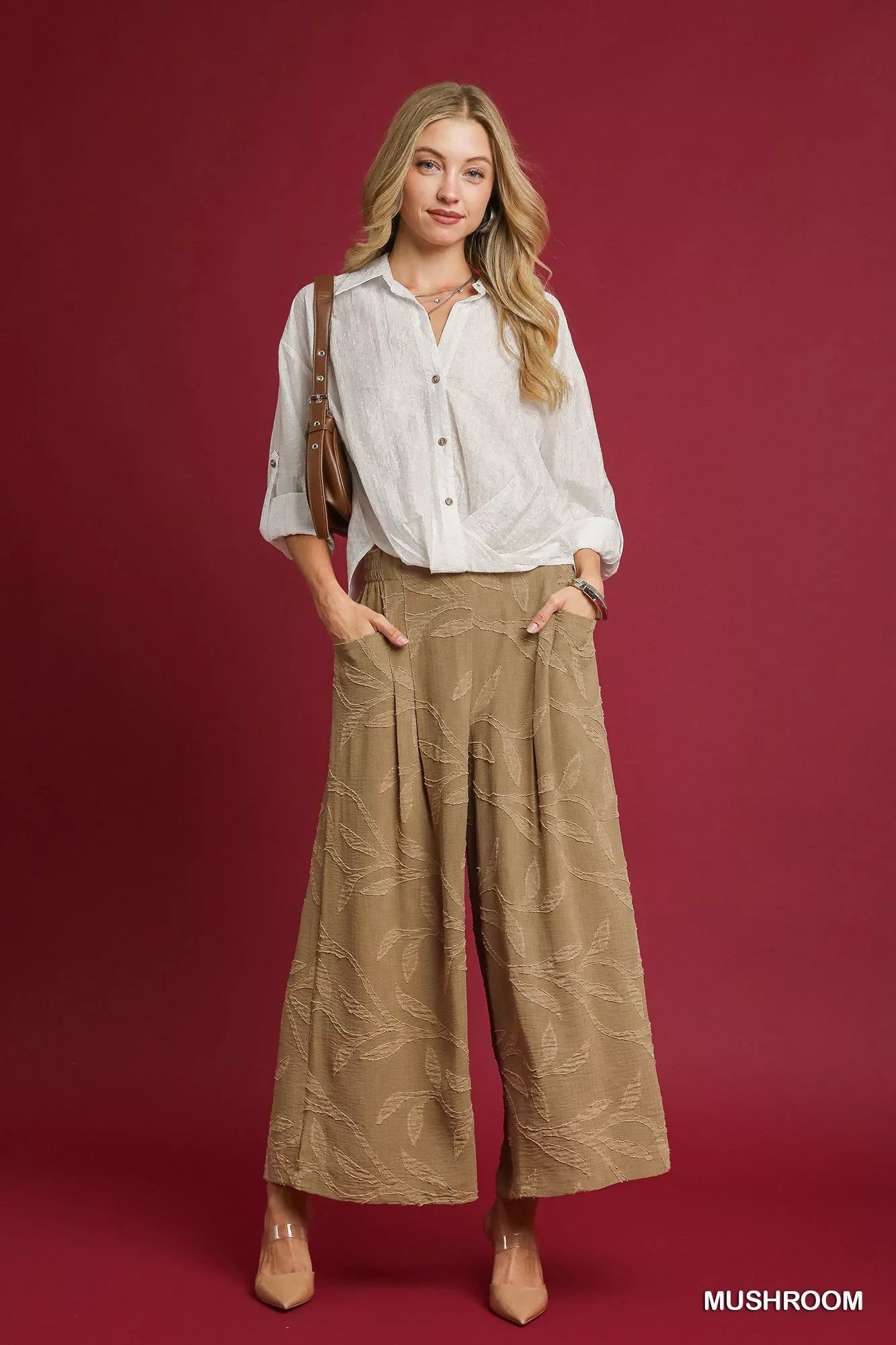 Model posing in jacquard wide-leg pants with intricate patterns, paired with a white button-up shirt and brown crossbody bag on red background.