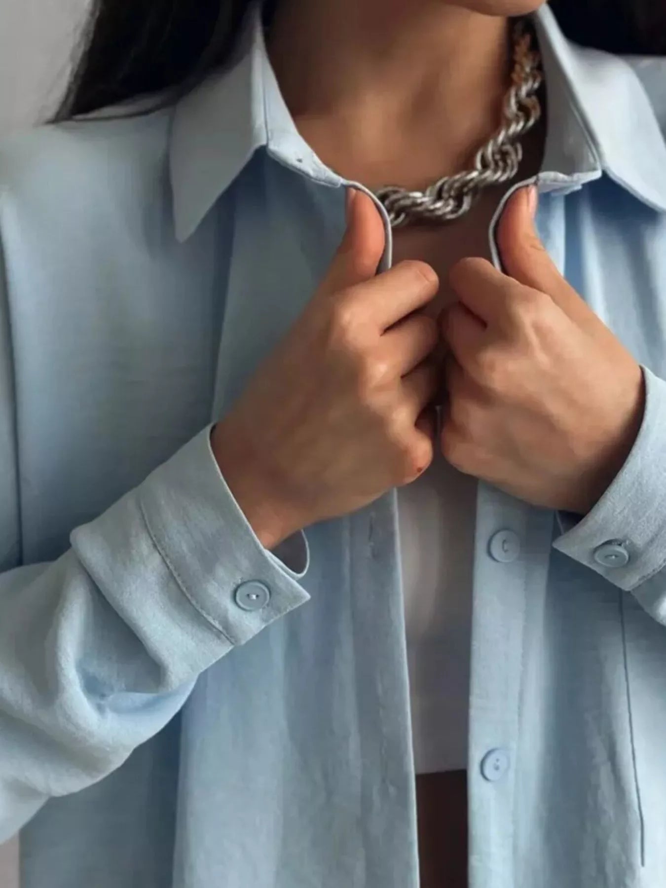 Woman wearing a light blue button-down shirt from a two-piece polyester elastane set, layered over a white tank top with silver chain necklace.
