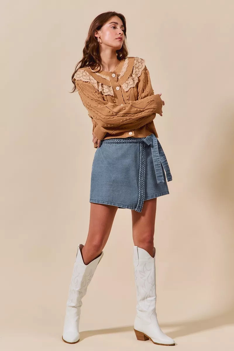 Woman modeling a wrap-style denim skort with braided trim, paired with a fringed sweater and white cowboy boots.