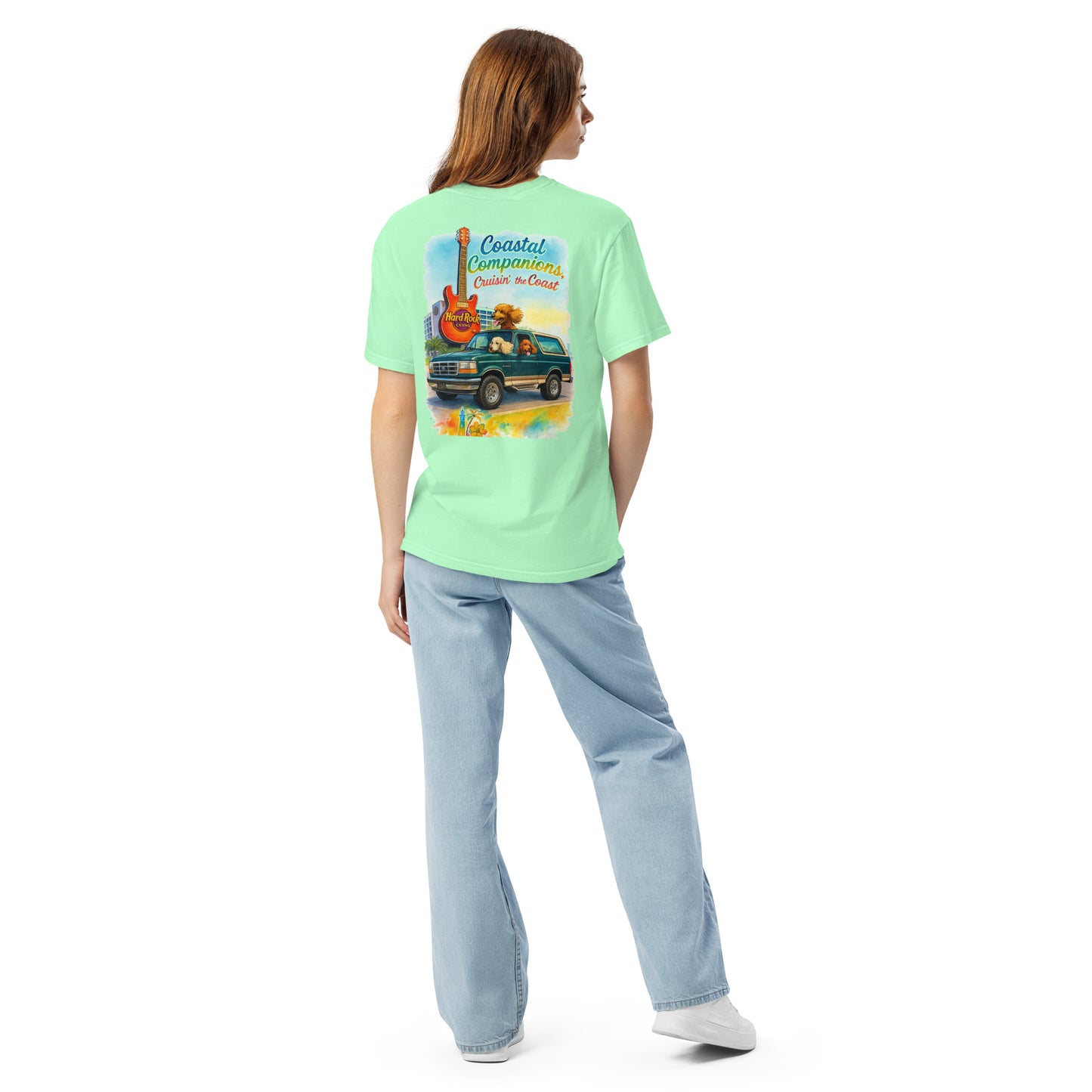 Back view of a woman wearing a light green short-sleeve tee with a coastal design featuring a guitar, truck, and dog.