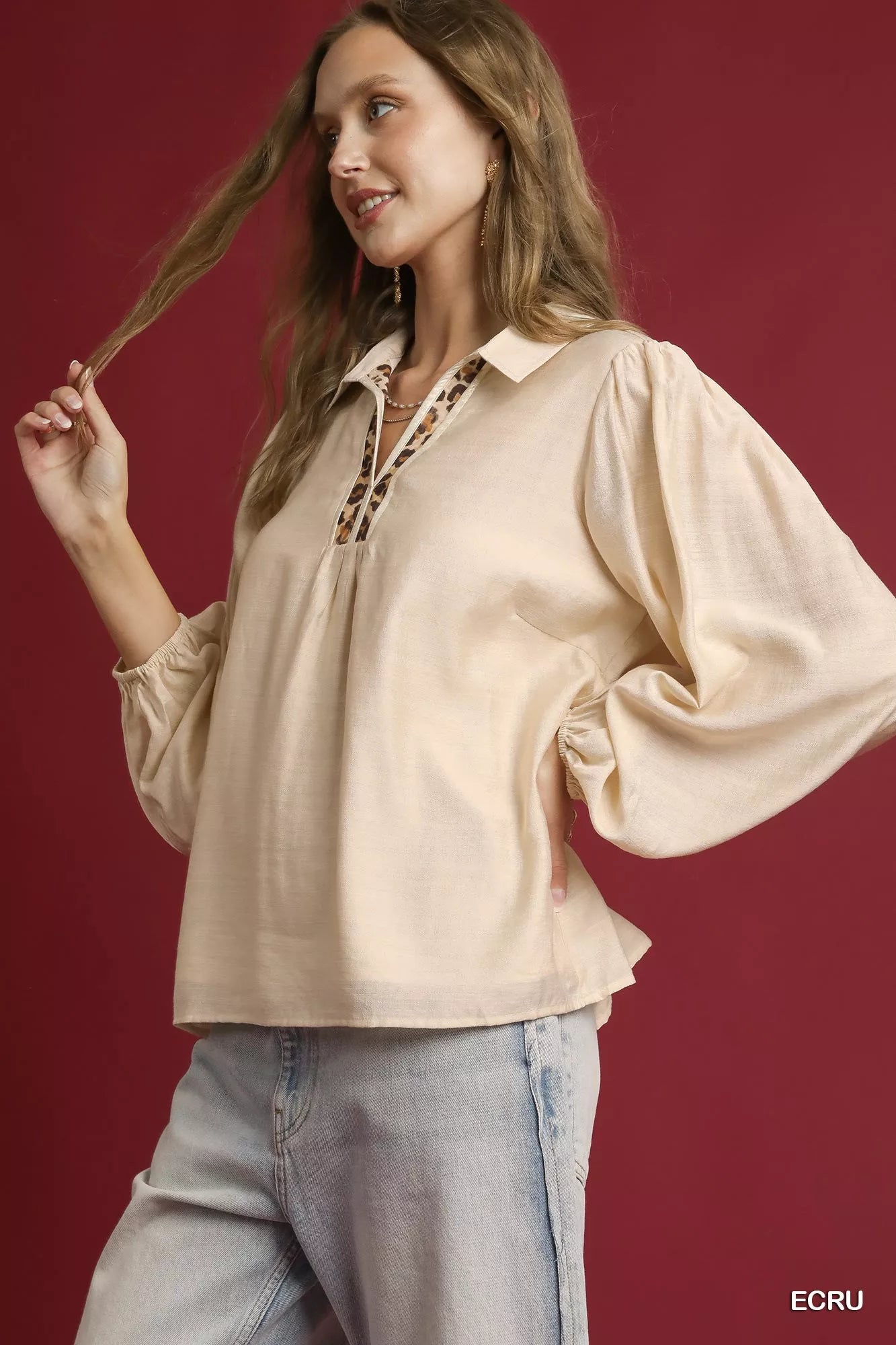 Woman modeling a long-sleeve beige blouse with leopard print trim on a red background.