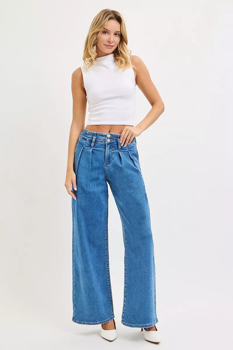 Model poses in RISEN mid-rise wide-leg pleated jeans, paired with a white tank top and white heels.
