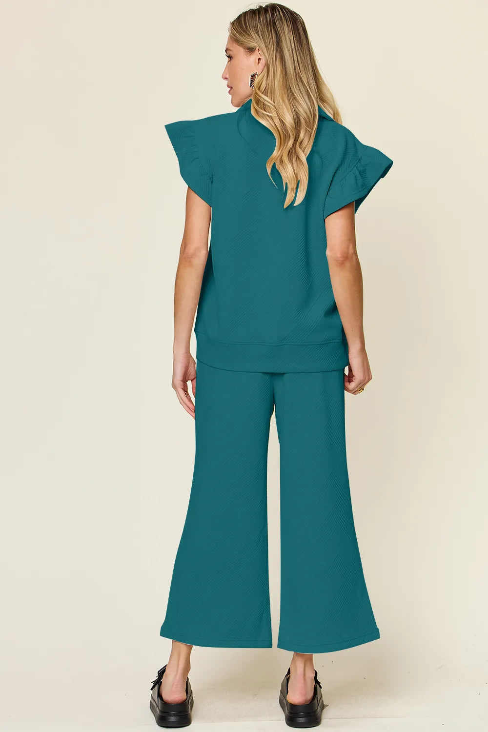 Rear view of a woman modeling a textured ruffle short sleeve top and wide leg pants set in teal.