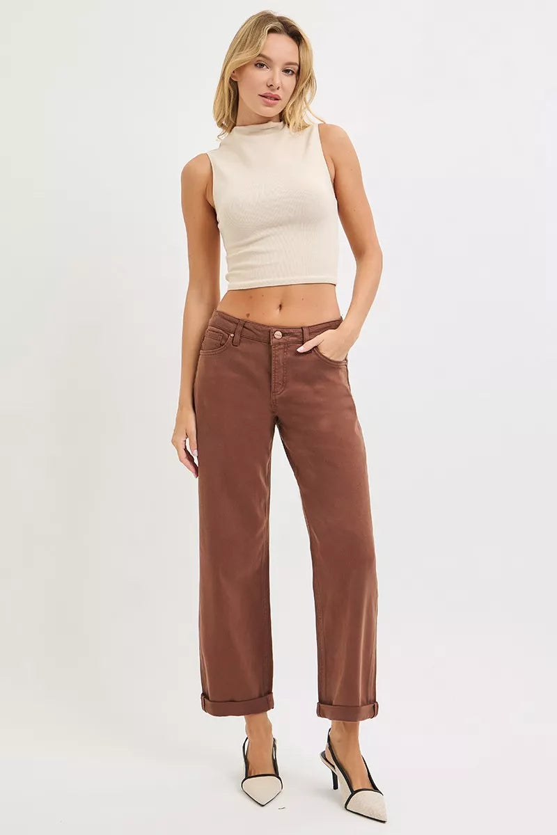 Model poses in brown wide-leg high-rise jeans with cuffed hems, paired with a beige mock-neck top and white heels.