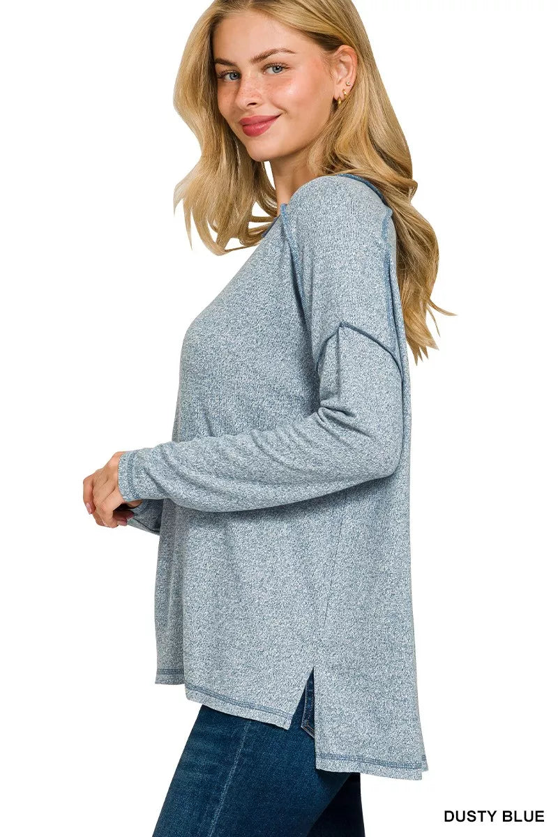 Woman modeling a dusty blue melange hacci hi-low tee with exposed seams and long sleeves.