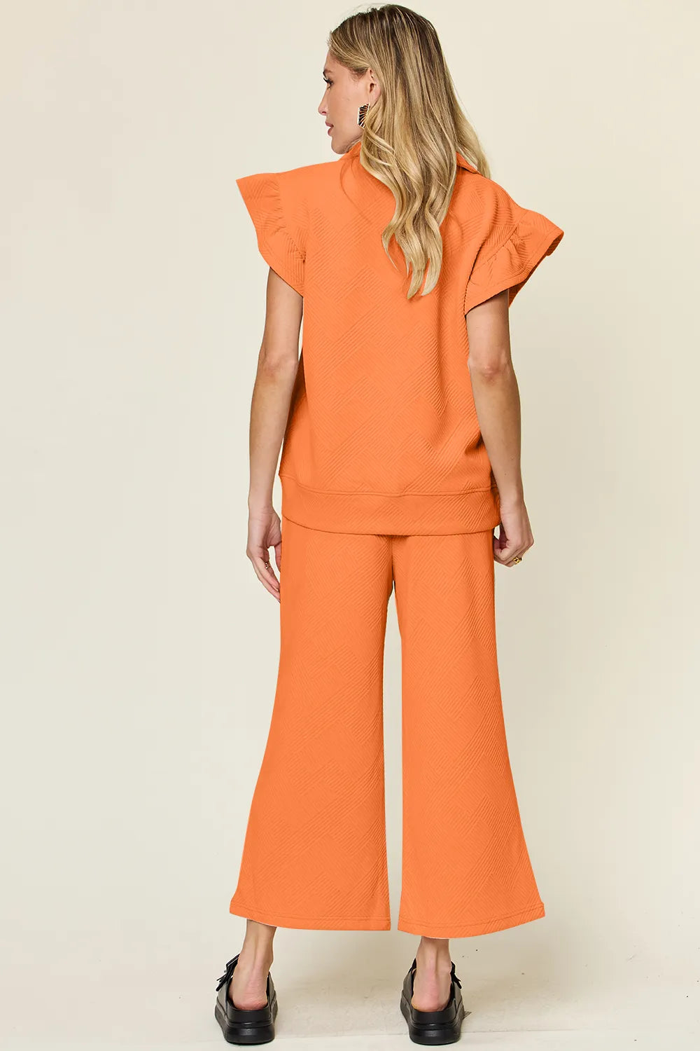 Rear view of a woman modeling a textured orange ruffle short sleeve top and wide leg pants set.