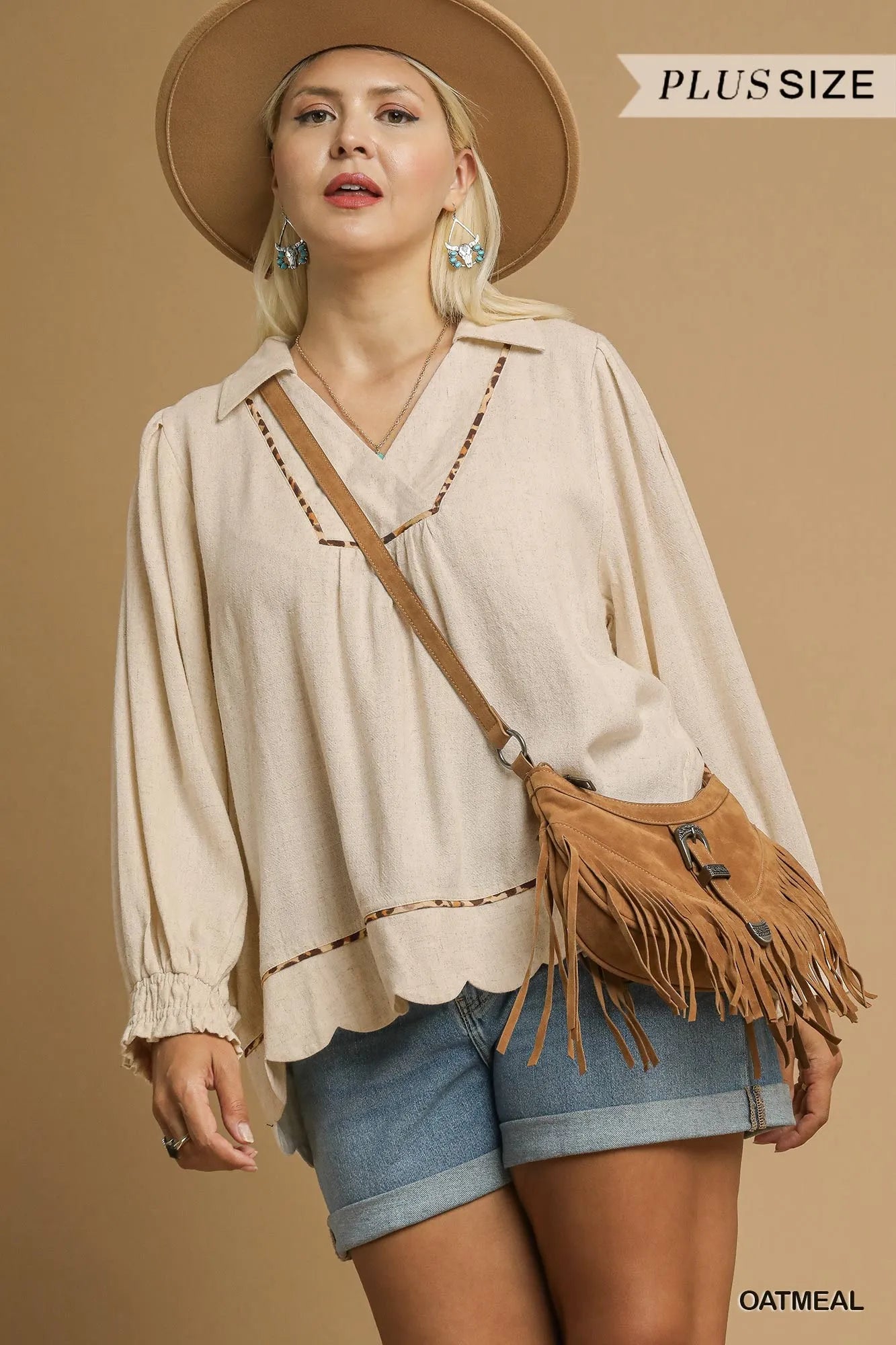 Plus-size woman modeling a beige linen scallop hem blouse with contrast trim, paired with denim shorts and a fringed bag.