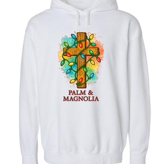 White hoodie with rustic wooden cross wrapped in colorful Christmas lights and "Palm & Magnolia" text.